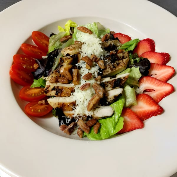 Grilled chicken salad with strawberries, cherry tomatoes, pecans, and shredded cheese on a white plate.